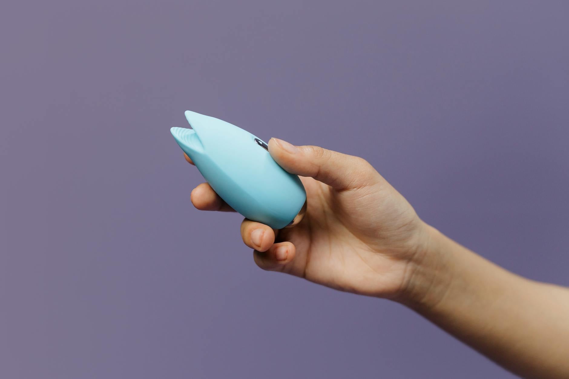 A blue silicone clitoral vibrator held in hand against a purple background, showing modern pleasure design